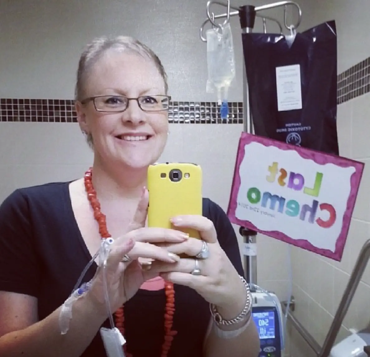 Person with an IV takes a mirror selfie beside infusion equipment and a colorful “Last Chemo” sign.
