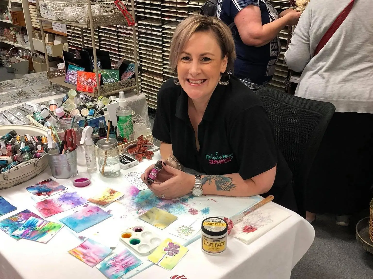 Smiling person sits at a craft table with inks, brushes, stencils and colorful handmade sample cards.