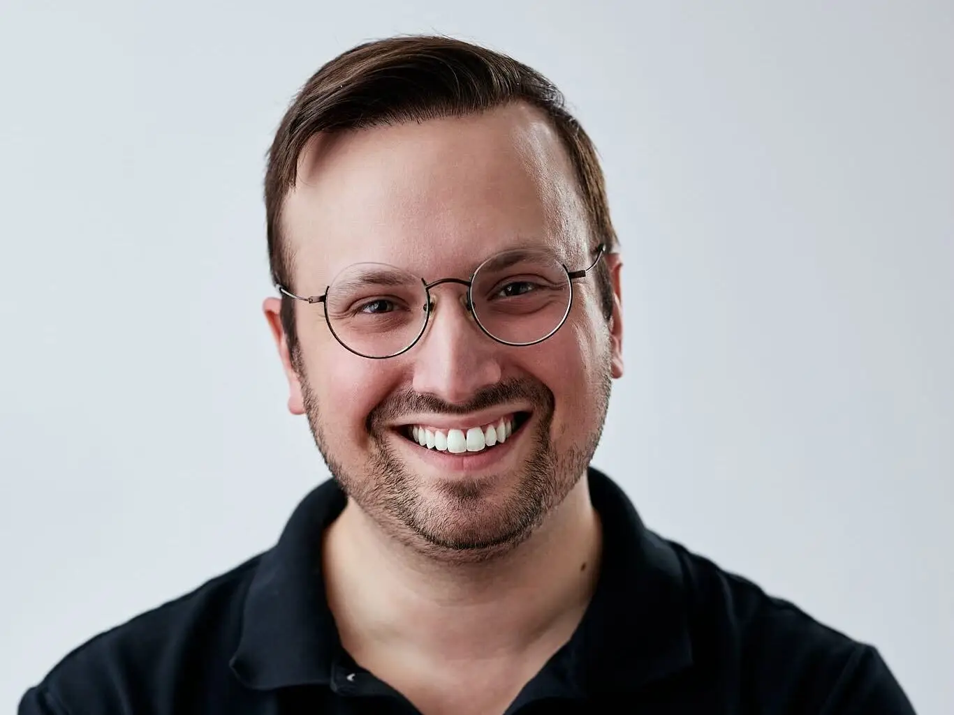 Smiling person wearing round glasses and a black polo stands with arms crossed against a light gray background.