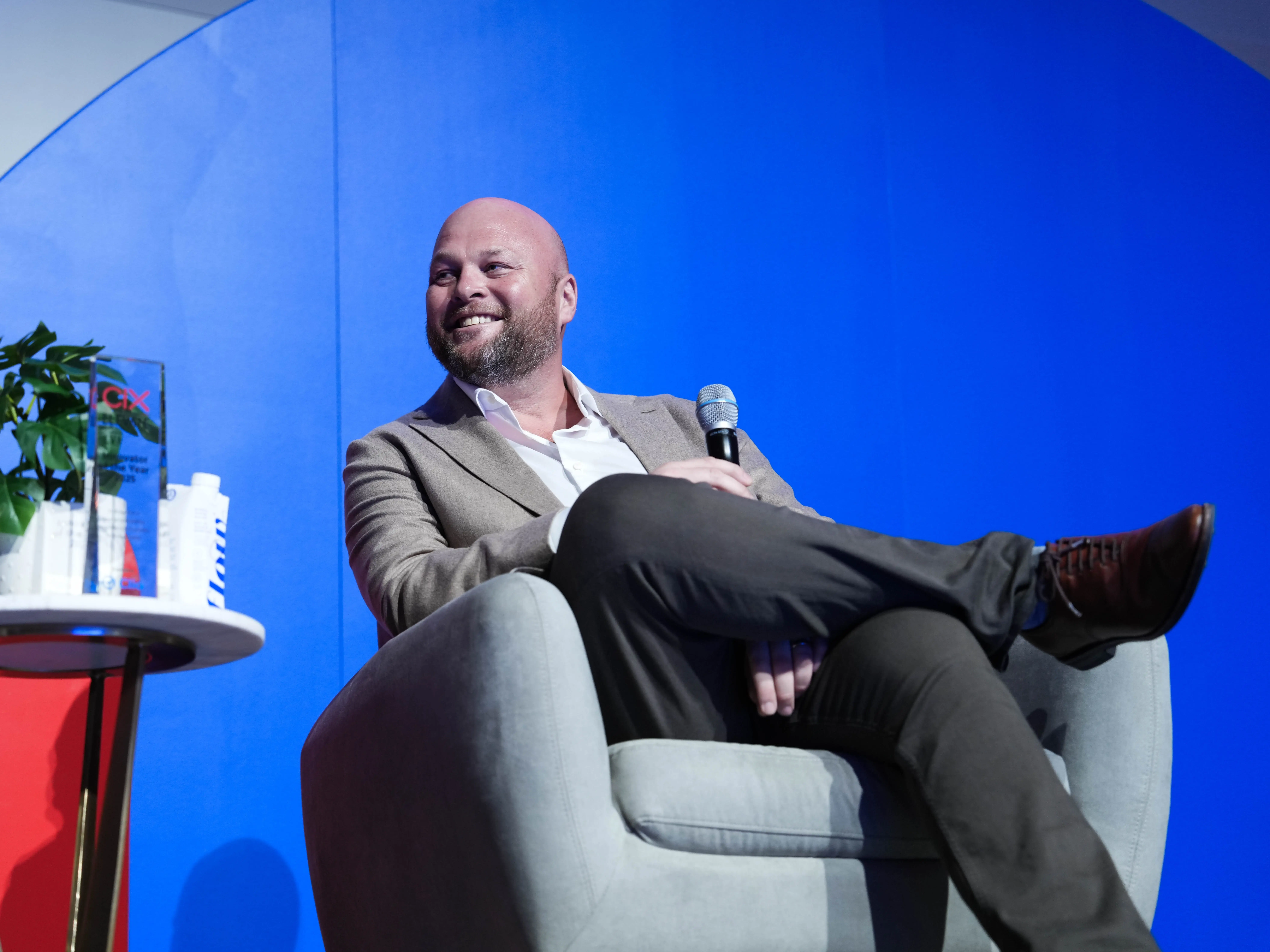 Speaker seated in an armchair holds a microphone on a blue-lit conference stage beside a small table.