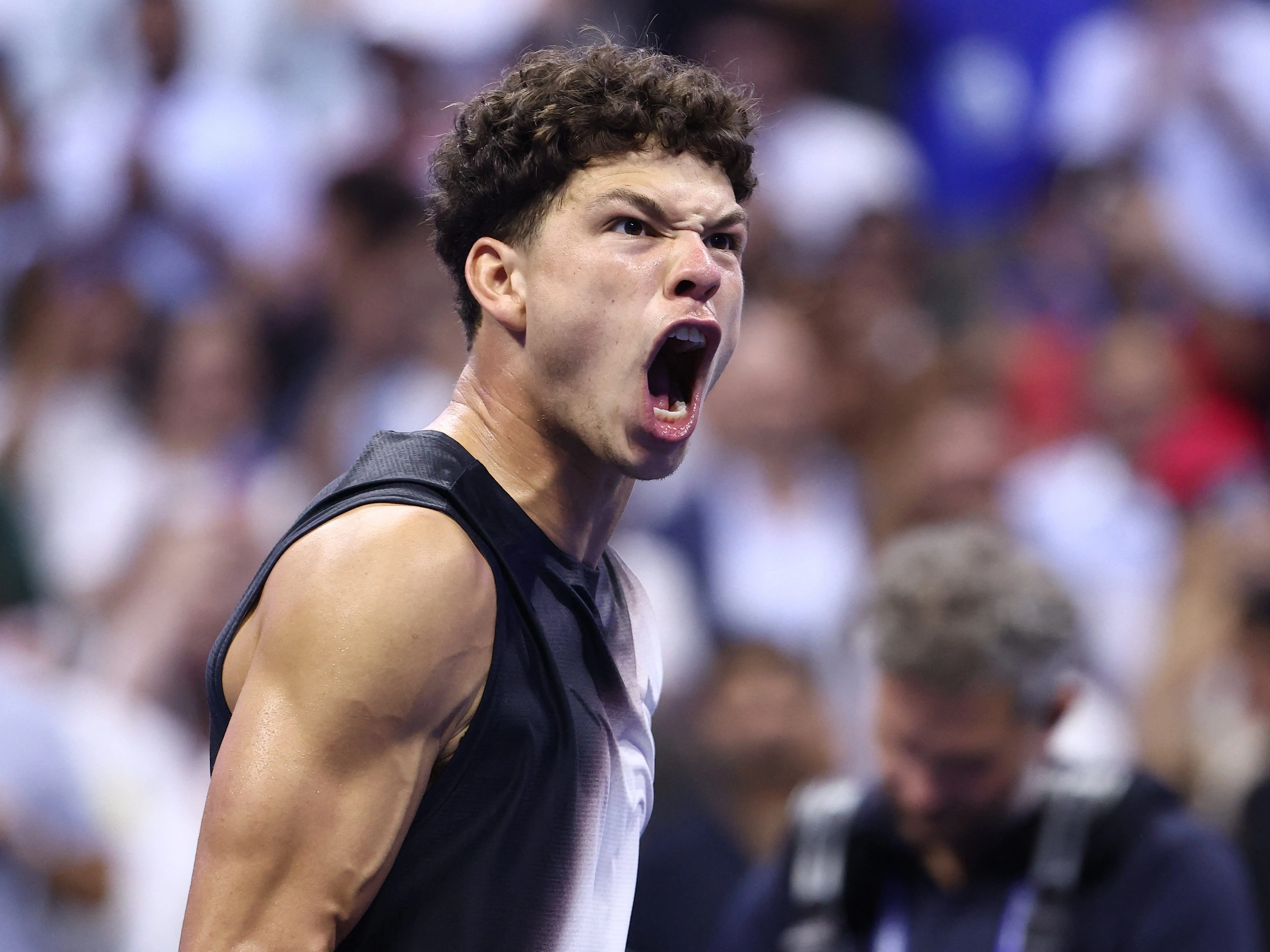 Ben Shelton shouts in celebration during a tennis match before a blurred crowd.