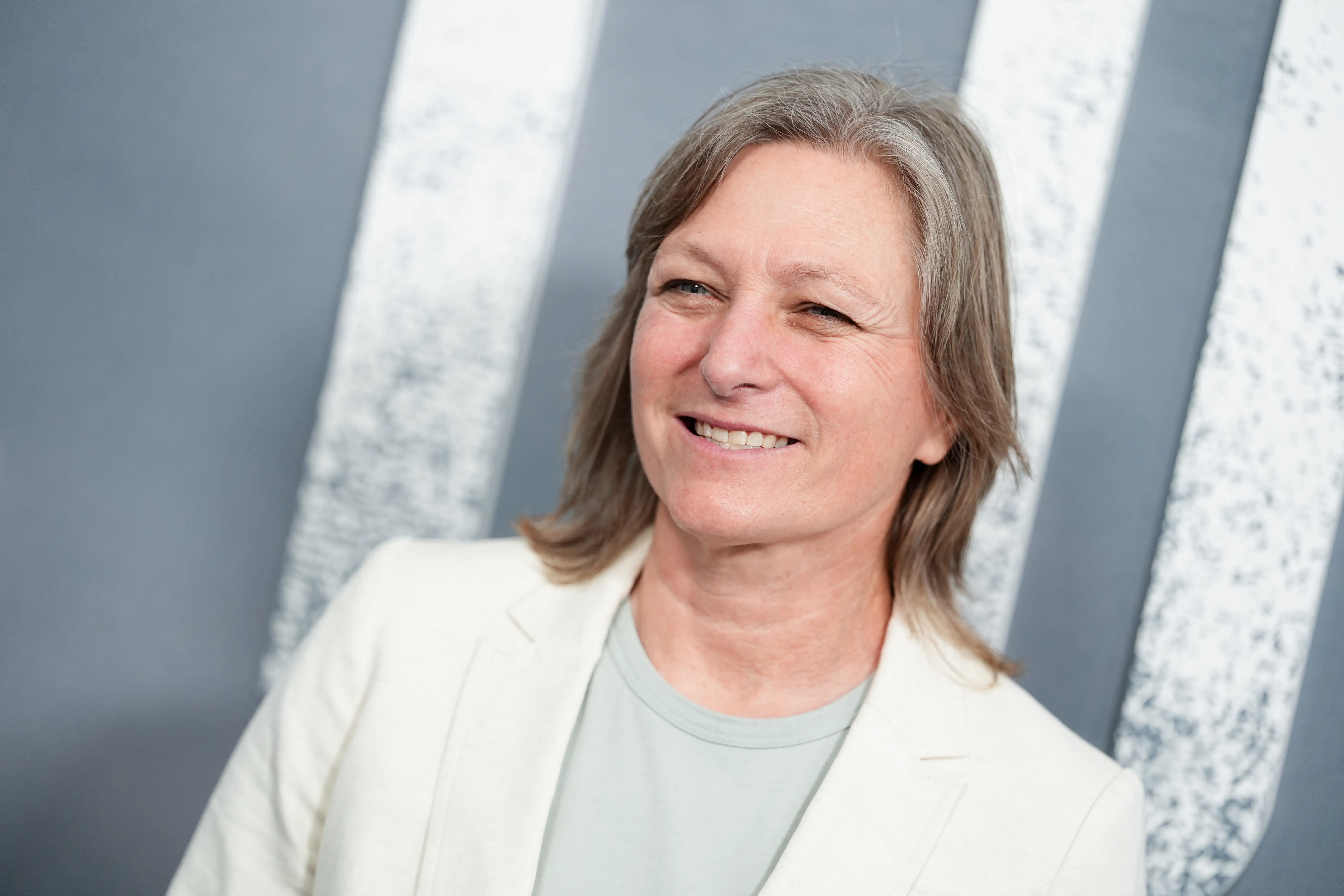 Cindy Holland with shoulder-length gray hair wears a white blazer in front of a striped gray backdrop.