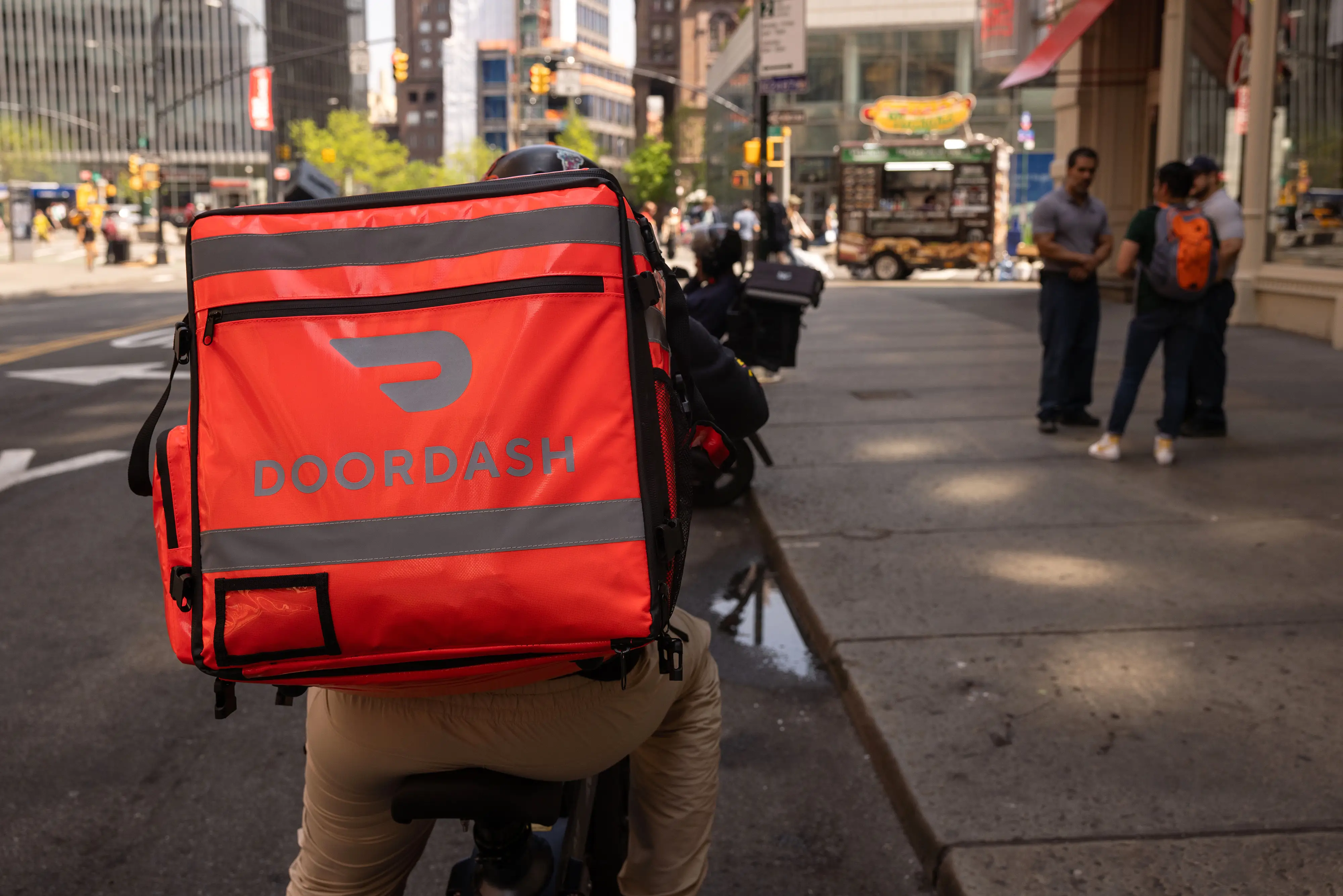 Cyclist carries a red DOORDASH delivery backpack on a busy city street with pedestrians and traffic lights.