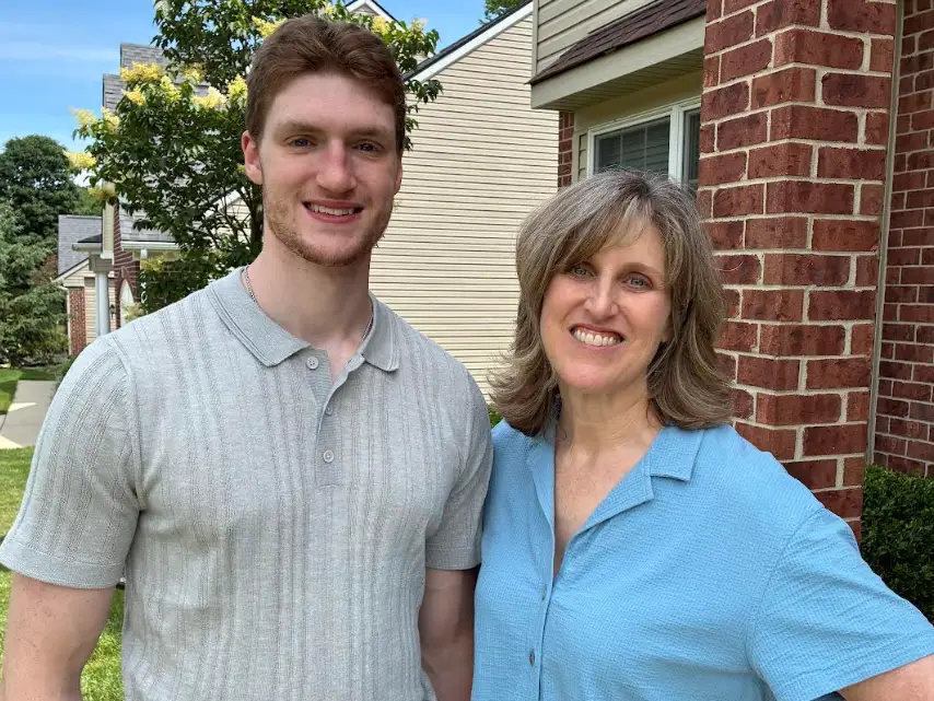 Two people smile outside a brick residential building on a sunny day.