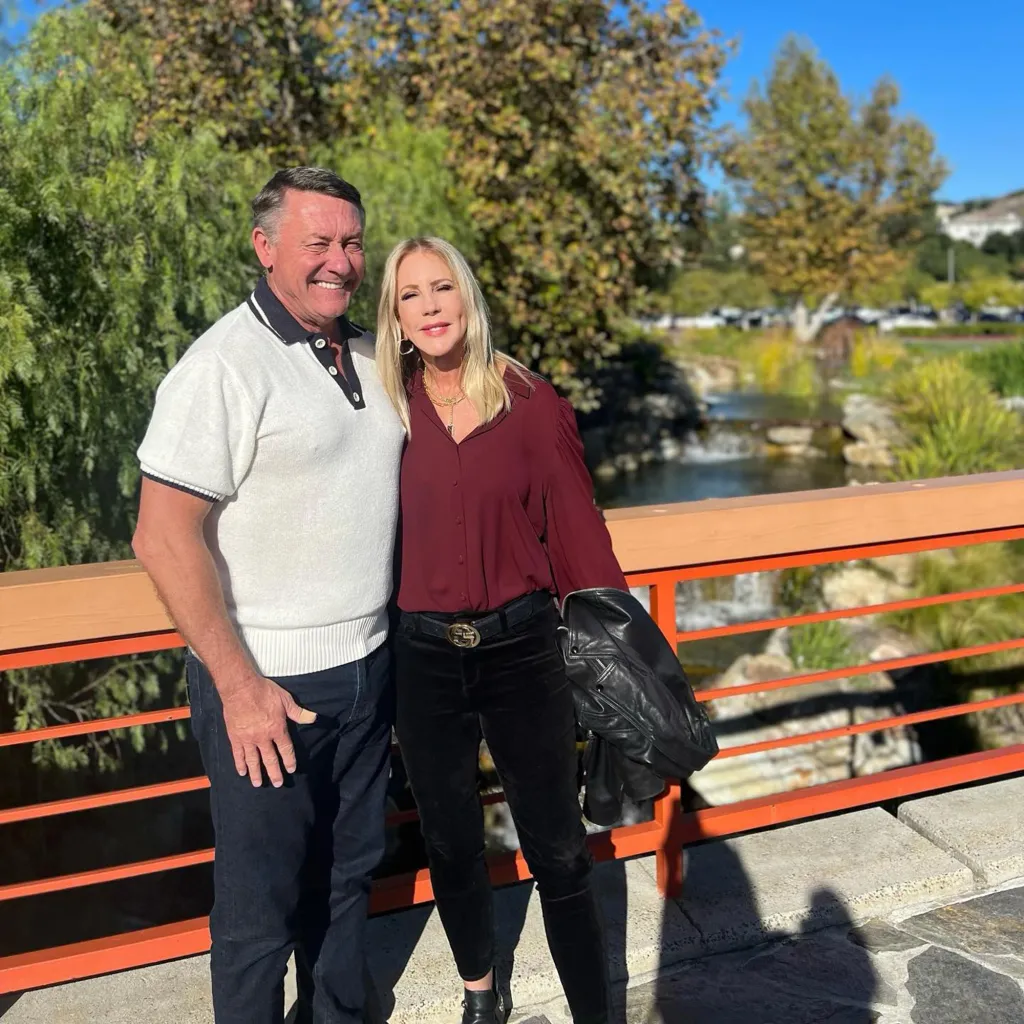 A man and a woman stand smiling on a bridge over a stream.