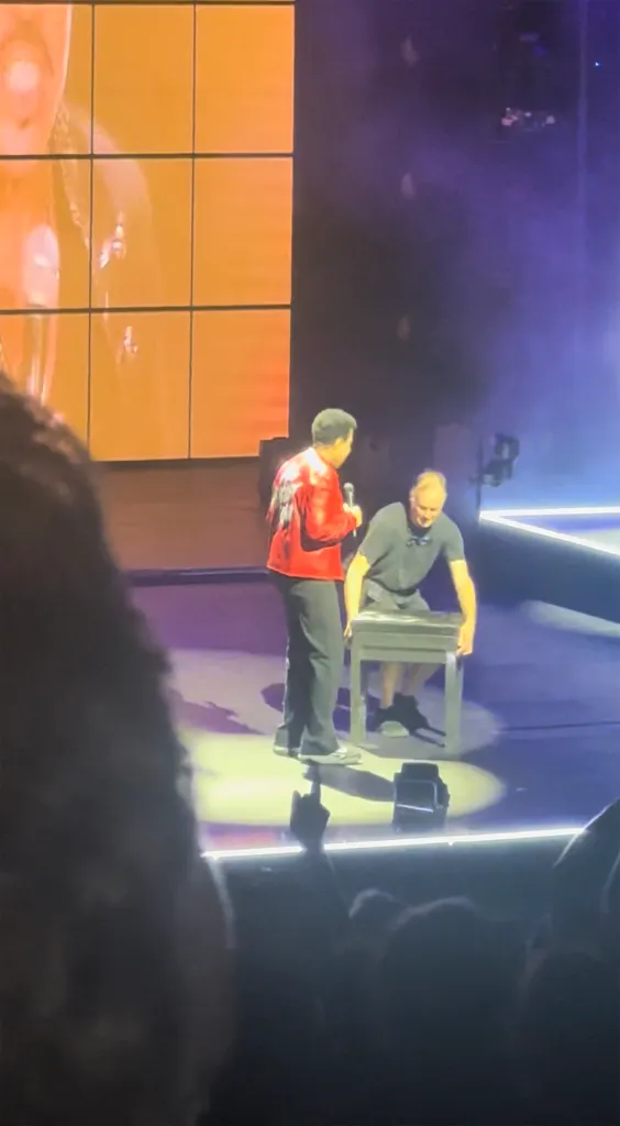 Lionel Richie, in a red shirt, speaks to an assistant who retrieves a stool for him on stage.