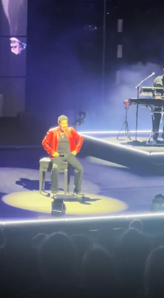Lionel Richie sitting on a stool on stage during a concert.
