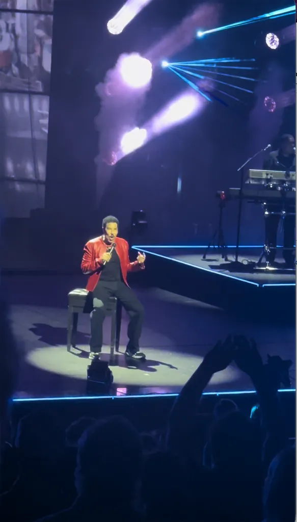 Lionel Richie seated on a stool, singing into a microphone on stage, with a band and audience visible.