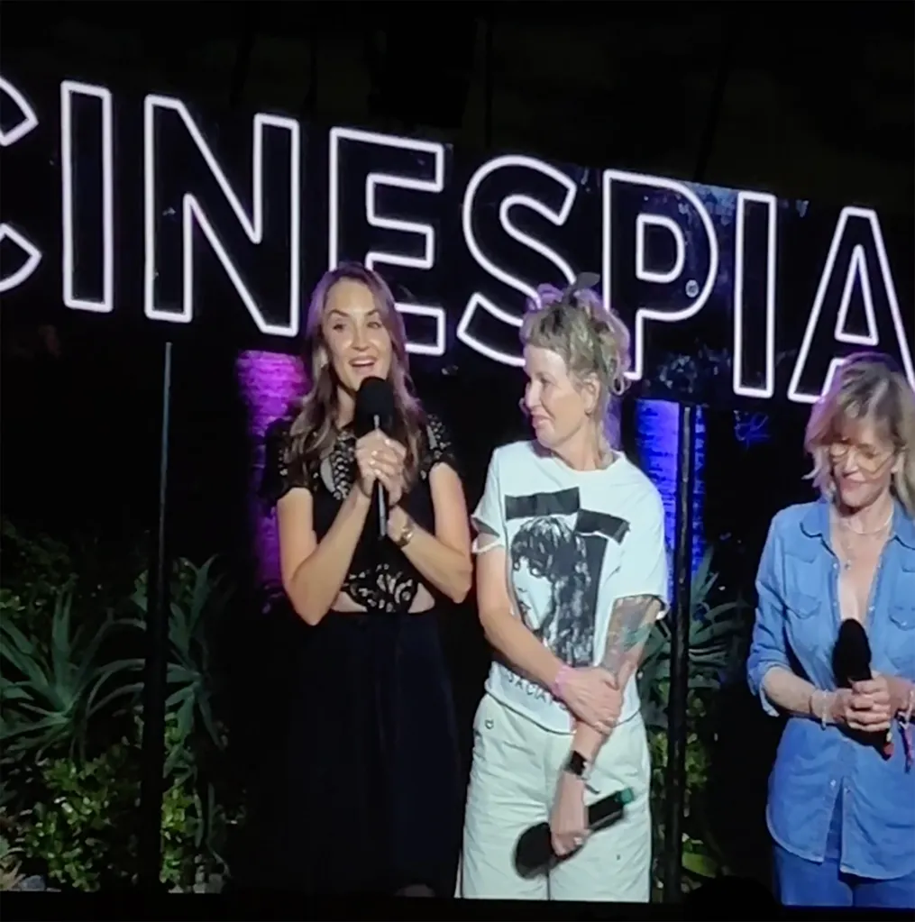 Rachael Leigh Cook and two other women holding microphones on stage in front of a neon sign.