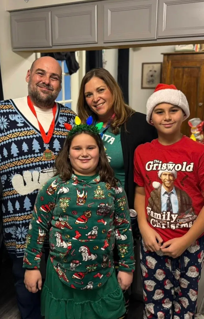 A family of four, including two adults and two children, smiling and wearing Christmas-themed clothing.