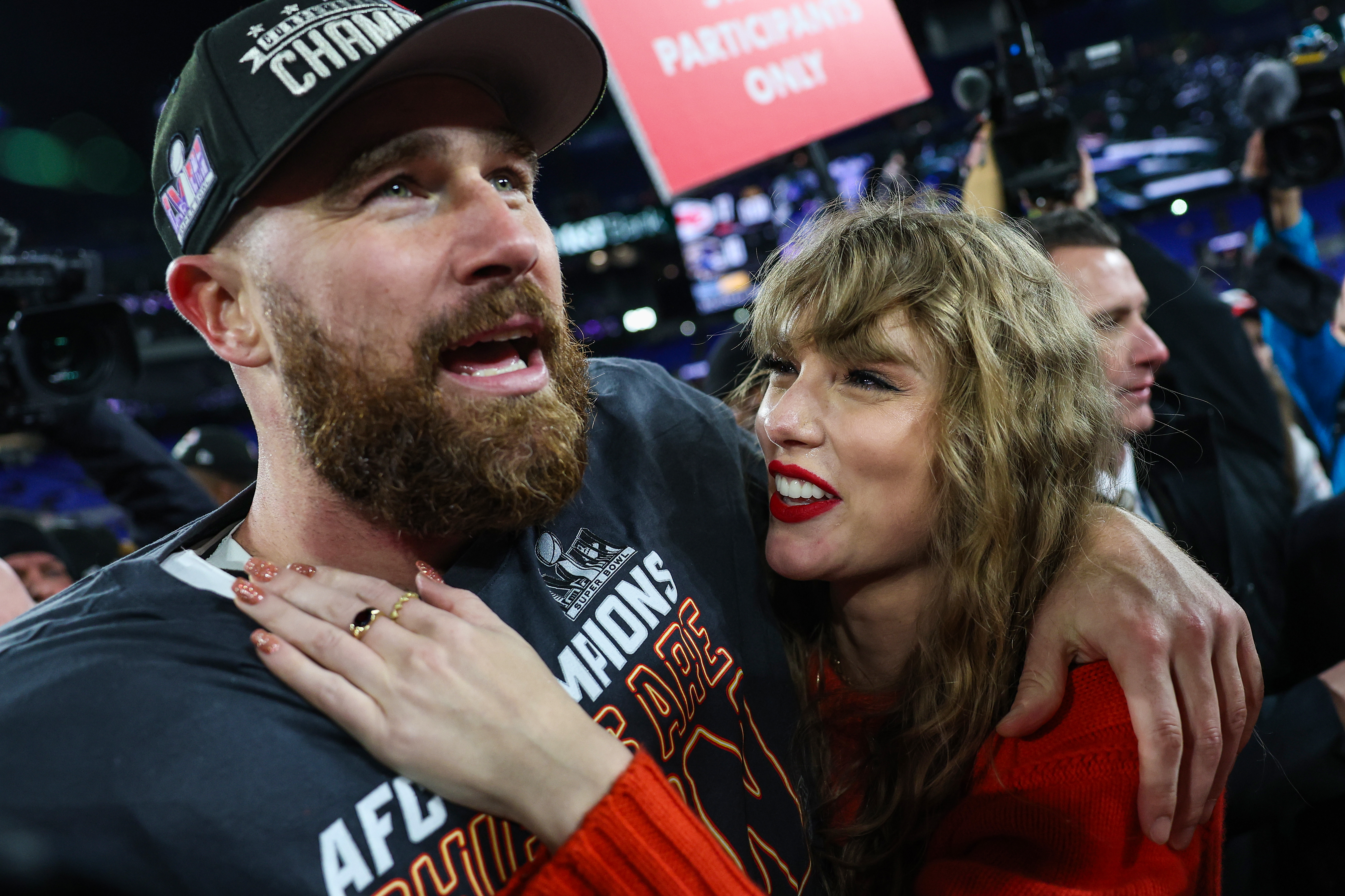 Travis Kelce, wearing a black Chiefs hat and shirt, celebrates with Taylor Swift, who wears a red sweater and has her arm around him.