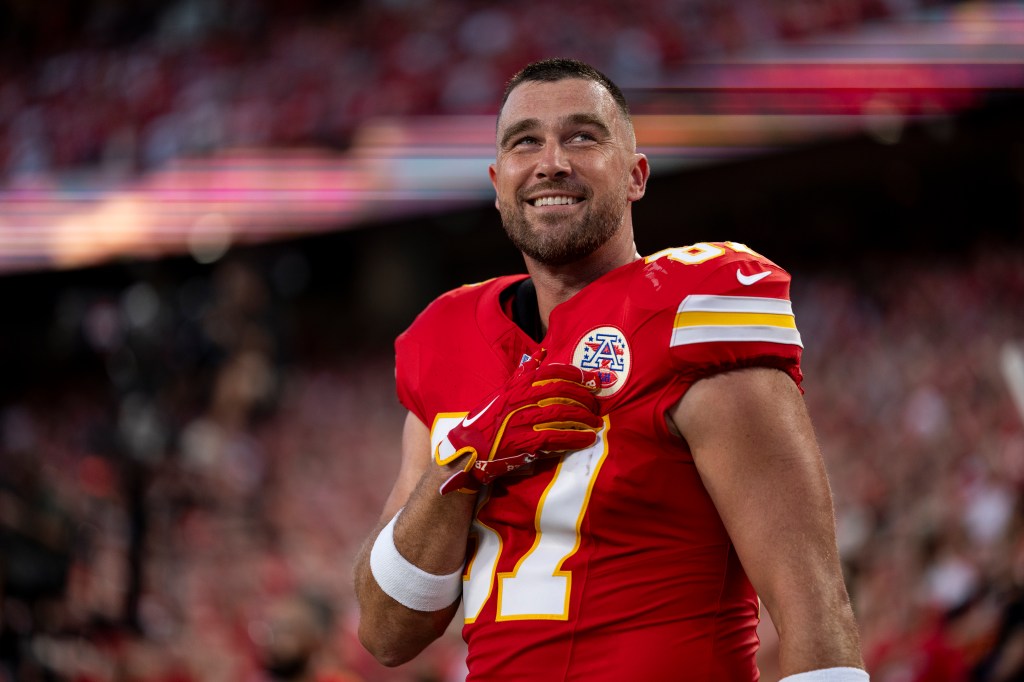 Travis Kelce, wearing a red Kansas City Chiefs uniform, smiles while looking up.