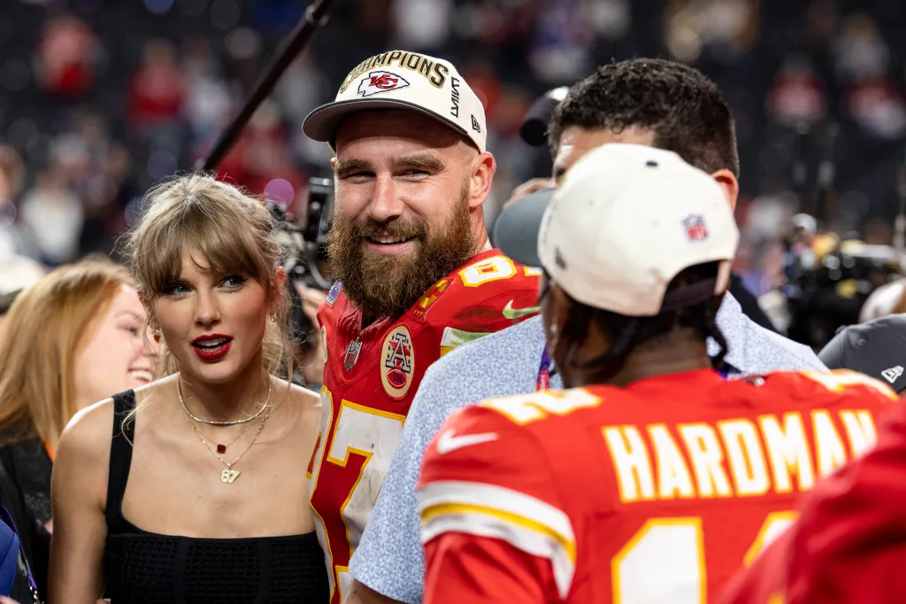 Travis Kelce, wearing a Kansas City Chiefs jersey and a Super Bowl LVIII Champions cap, smiles next to Taylor Swift while they talk to Mecole Hardman Jr. (seen from behind) on the field after the Super Bowl.