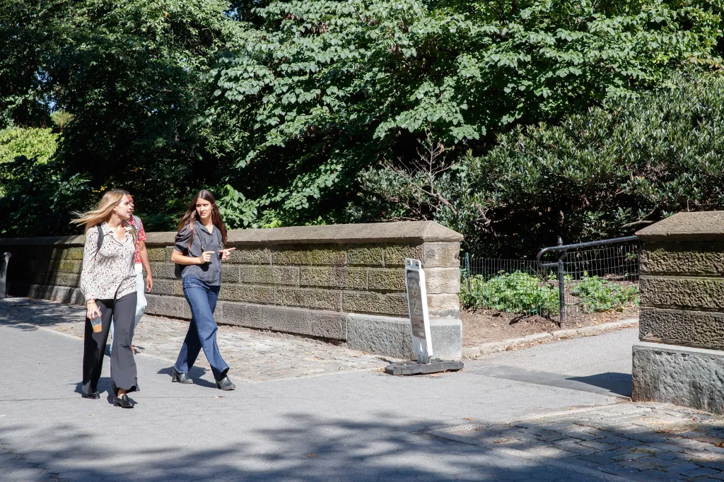 Three people walk along a paved path next to a stone wall in Central Park.