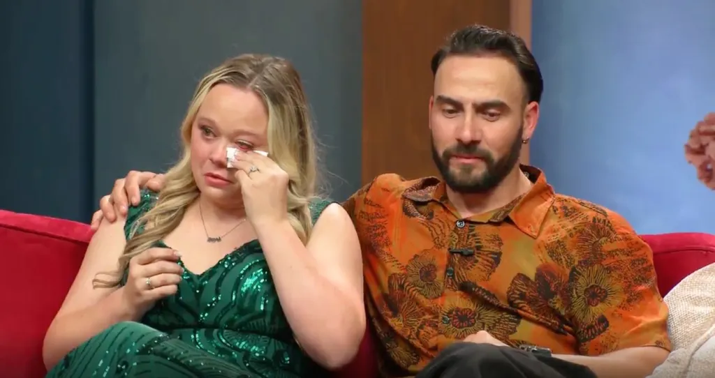 A woman in a green sequined dress wipes her crying eyes with a tissue, while a man with a beard sits beside her with his arm around her.