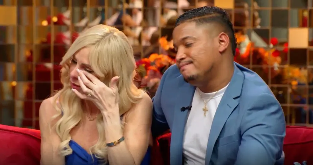 A blonde woman in a blue dress cries while a man in a blue blazer and white t-shirt looks on sympathetically.