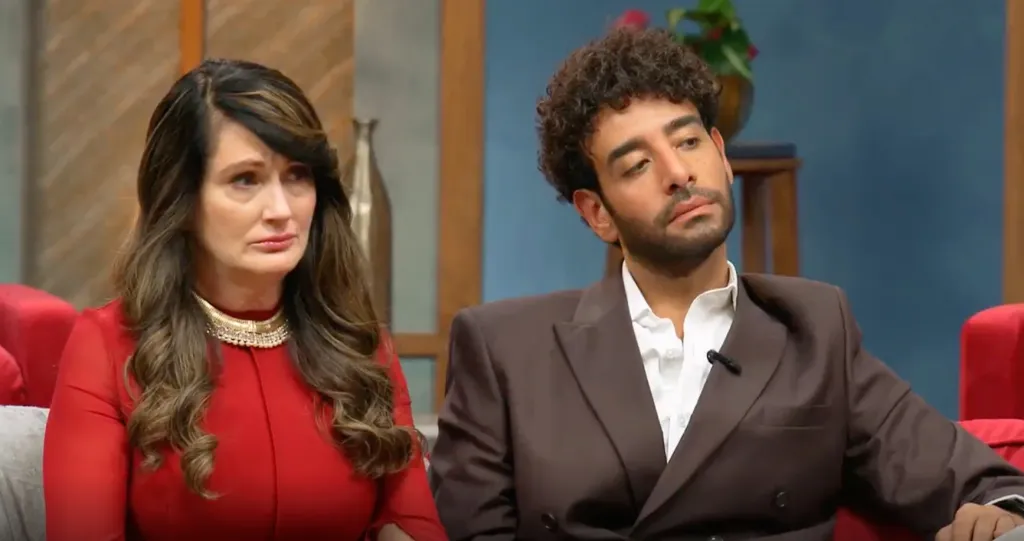 A woman in a red dress with a gold necklace and a man in a brown suit looking upset.