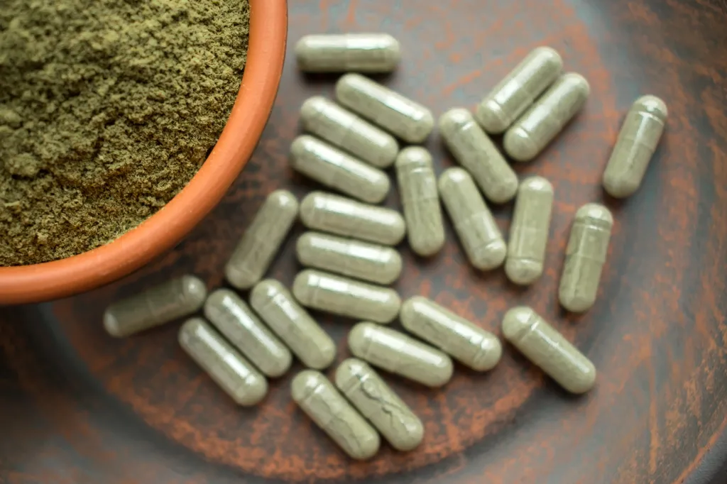 Kratom powder in an orange bowl and several kratom capsules scattered on a brown plate.
