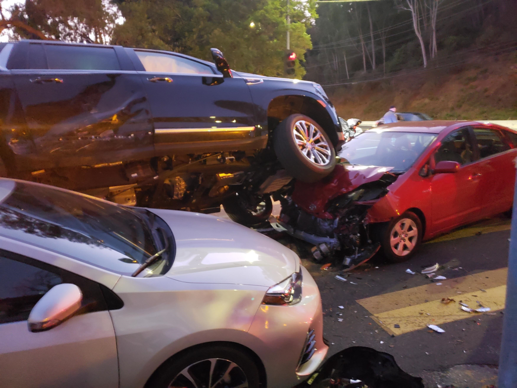 A multicar accident involving Arnold Schwarzenegger's dark SUV resting on top of a red car, with a white car nearby.