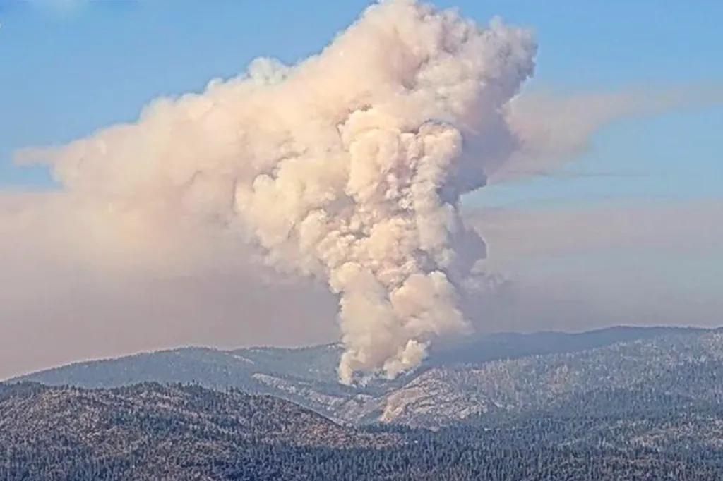 A large column of smoke rising from a wildfire in a forested mountain range.