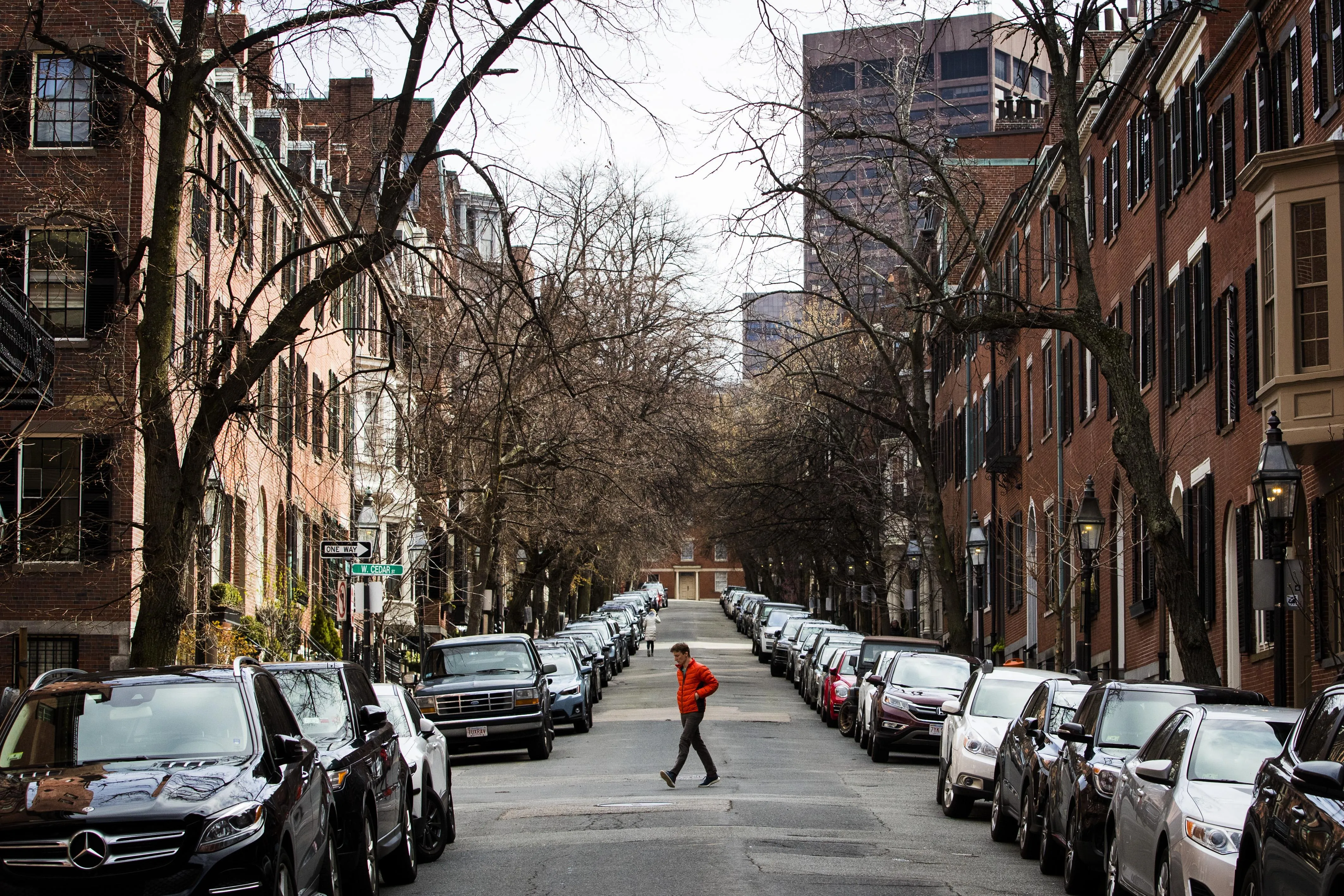 A person in a bright orange jacket crosses a narrow residential street lined with parked cars, leafless trees, and historic brick townhouses, with taller city buildings visible in the background. A person in a bright orange jacket crosses a narrow residential street lined with parked cars, leafless trees, and historic brick townhouses, with taller city buildings visible in the background.