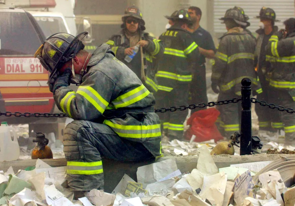 Firefighter sitting on the ground with head in hands, surrounded by debris, with other firefighters in background.