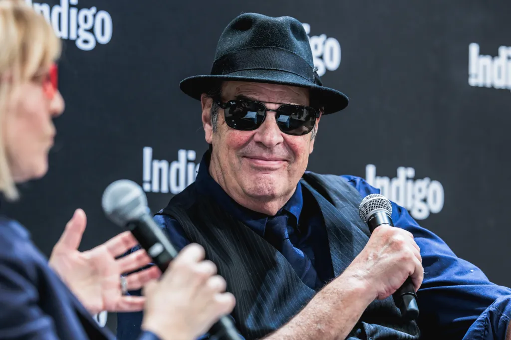 Dan Aykroyd, wearing a hat and sunglasses, holding a microphone.