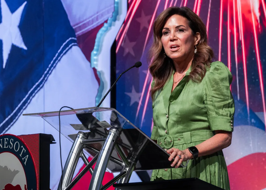 A woman in a green dress speaking at a podium with an American flag and a red-striped background.
