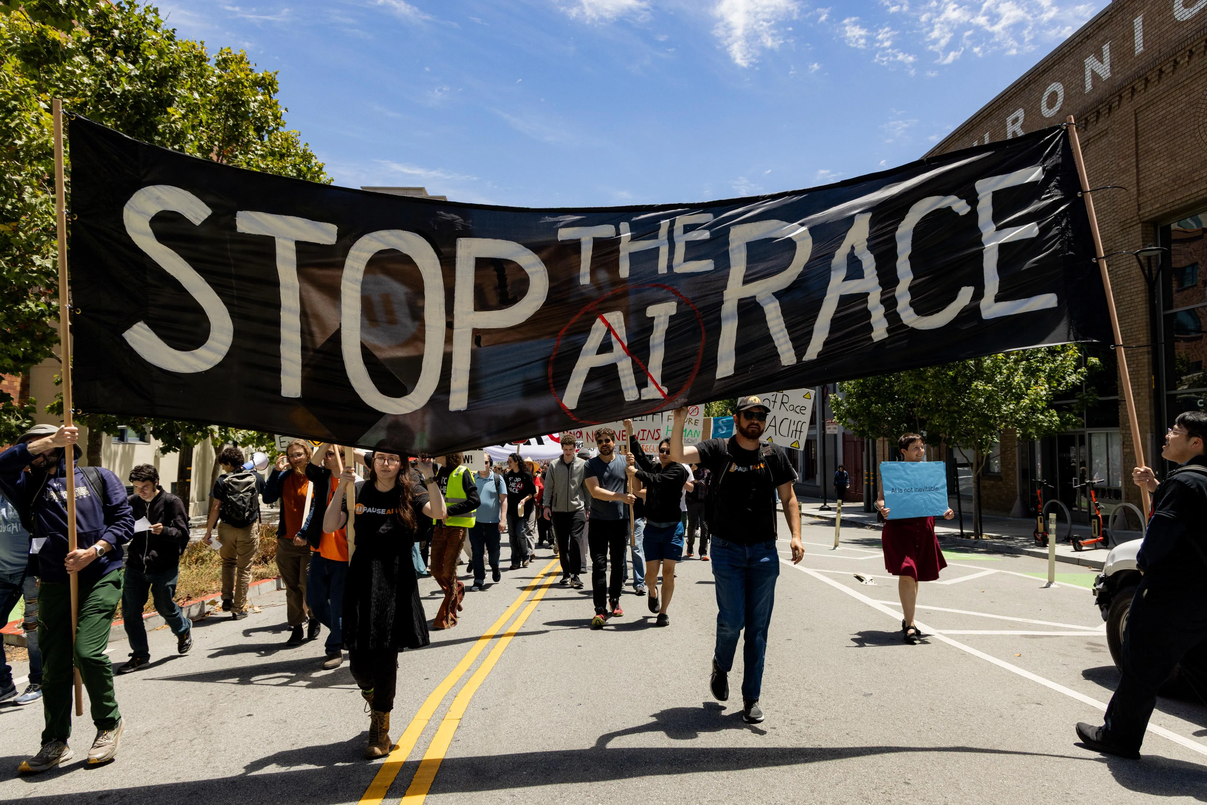 Protesters march down a sunny city street beneath a large black banner reading “STOP THE AI RACE,” with the letters “AI” crossed out in red.
