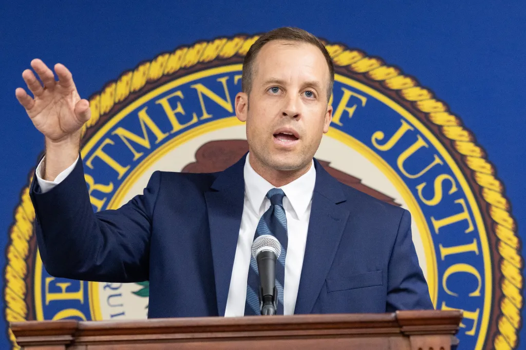 Man in a suit speaks at a podium with a microphone, gesturing with his right hand in front of a Department of Justice seal.