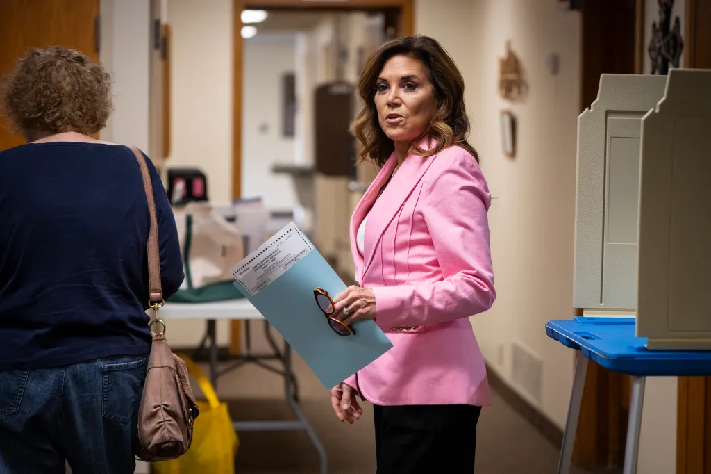 A woman in a pink blazer holds a blue folder and a Minnesota primary ballot from August 11, 2026.