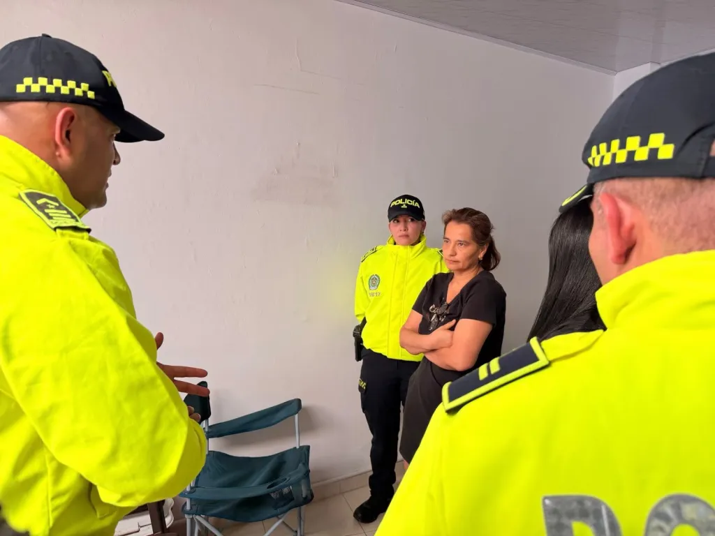 Two police officers in neon yellow uniforms and a woman with folded arms talking in a room.