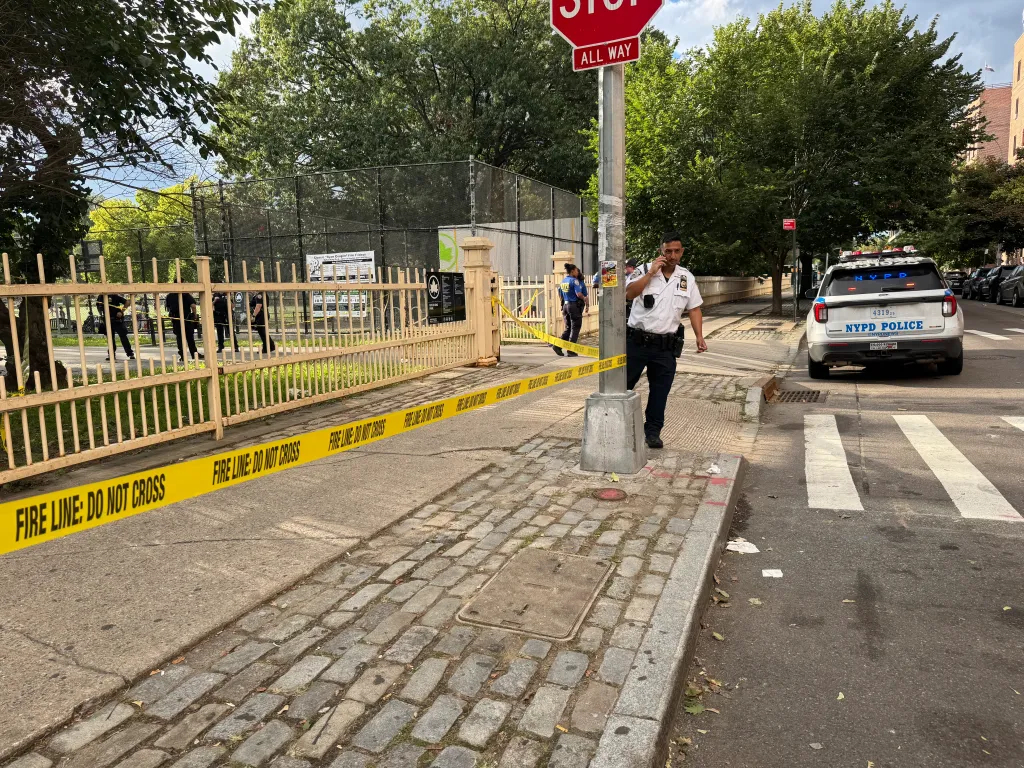 A police officer talking on a phone stands next to a fire line across a park entrance, with an NYPD vehicle parked nearby.