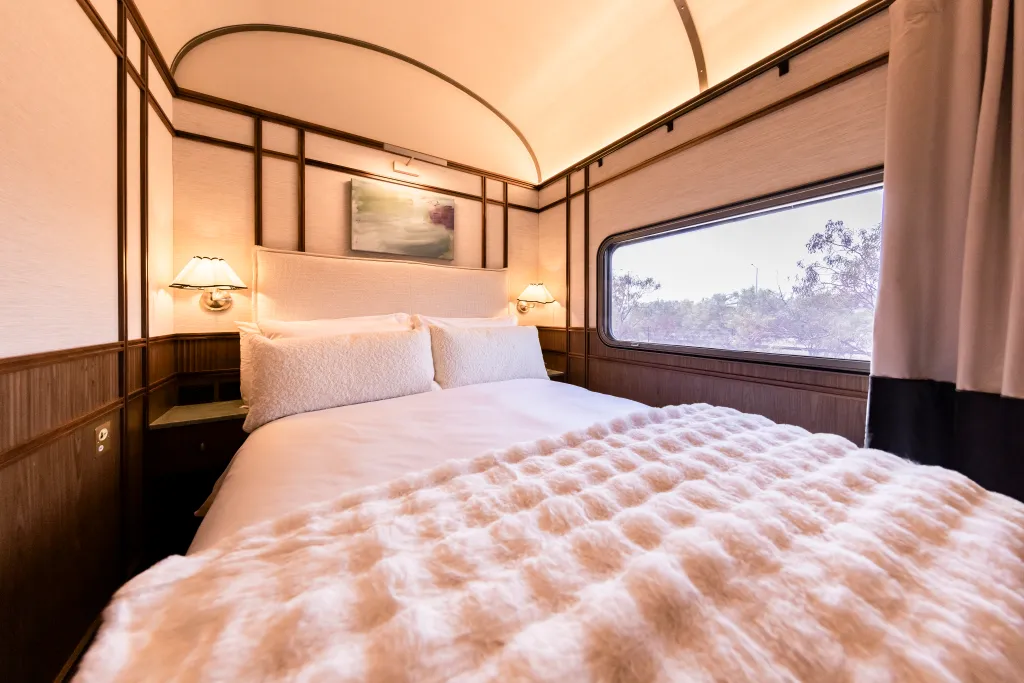 A bed with a white comforter and fluffy blanket, flanked by wall-mounted lamps, inside a luxury train suite.