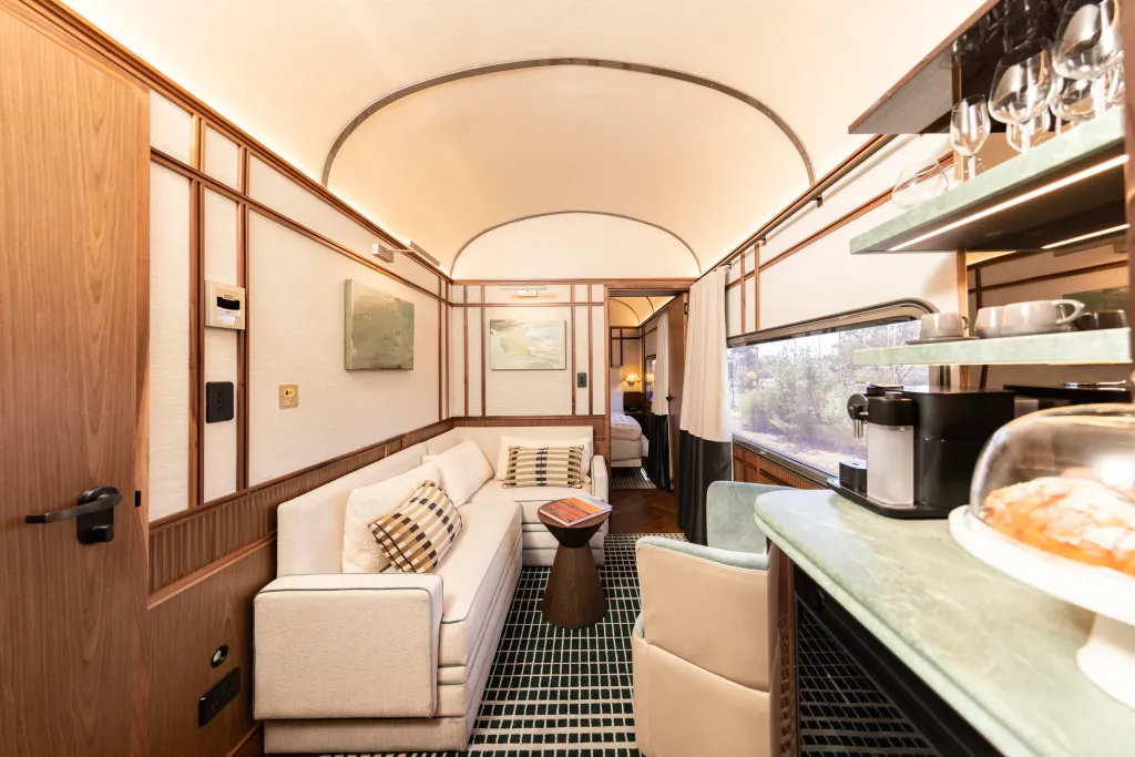 A living area in the Aurora or Austalis Suites, with a cream-colored sofa and green tiled rug, a small round table, and a built-in shelf with wine glasses, a coffee maker, and pastries, next to a window overlooking greenery.