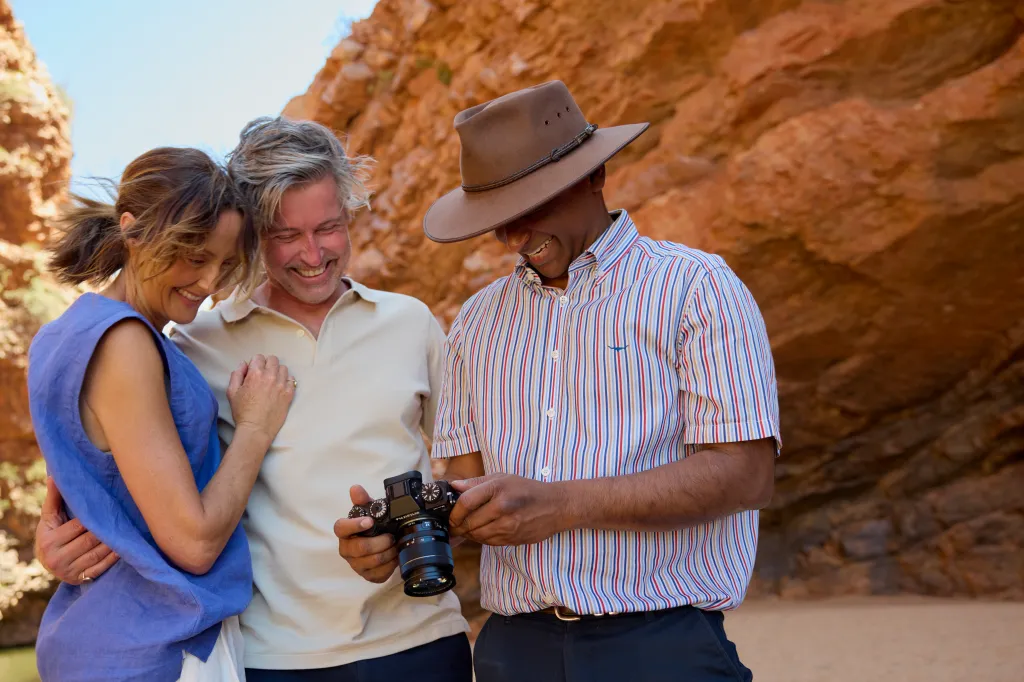 Three people looking at photos on a camera, smiling, with a rocky red landscape in the background.