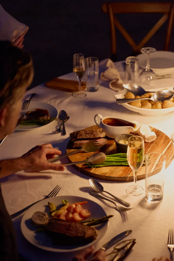 A person serves sliced beef from a wooden board at a dinner table, with side dishes and drinks.