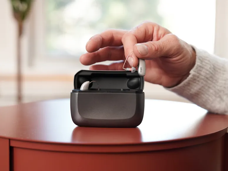 Grey rectangular case on wooden surface holding silver hearing aids with a one hand pulling a hearing aid out