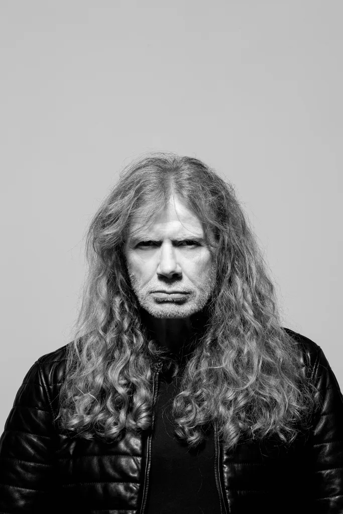Black and white photo of a man with long, curly hair and a beard, wearing a leather jacket, looking intensely at the camera.