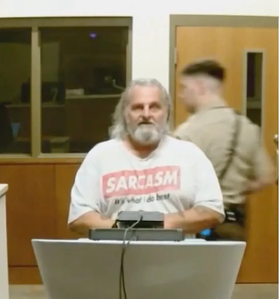 A man with long gray hair and a beard sits at a table wearing a white t-shirt that says 