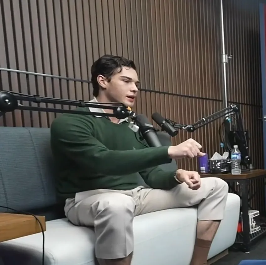 Man in a green sweater and khaki pants speaking into a microphone.