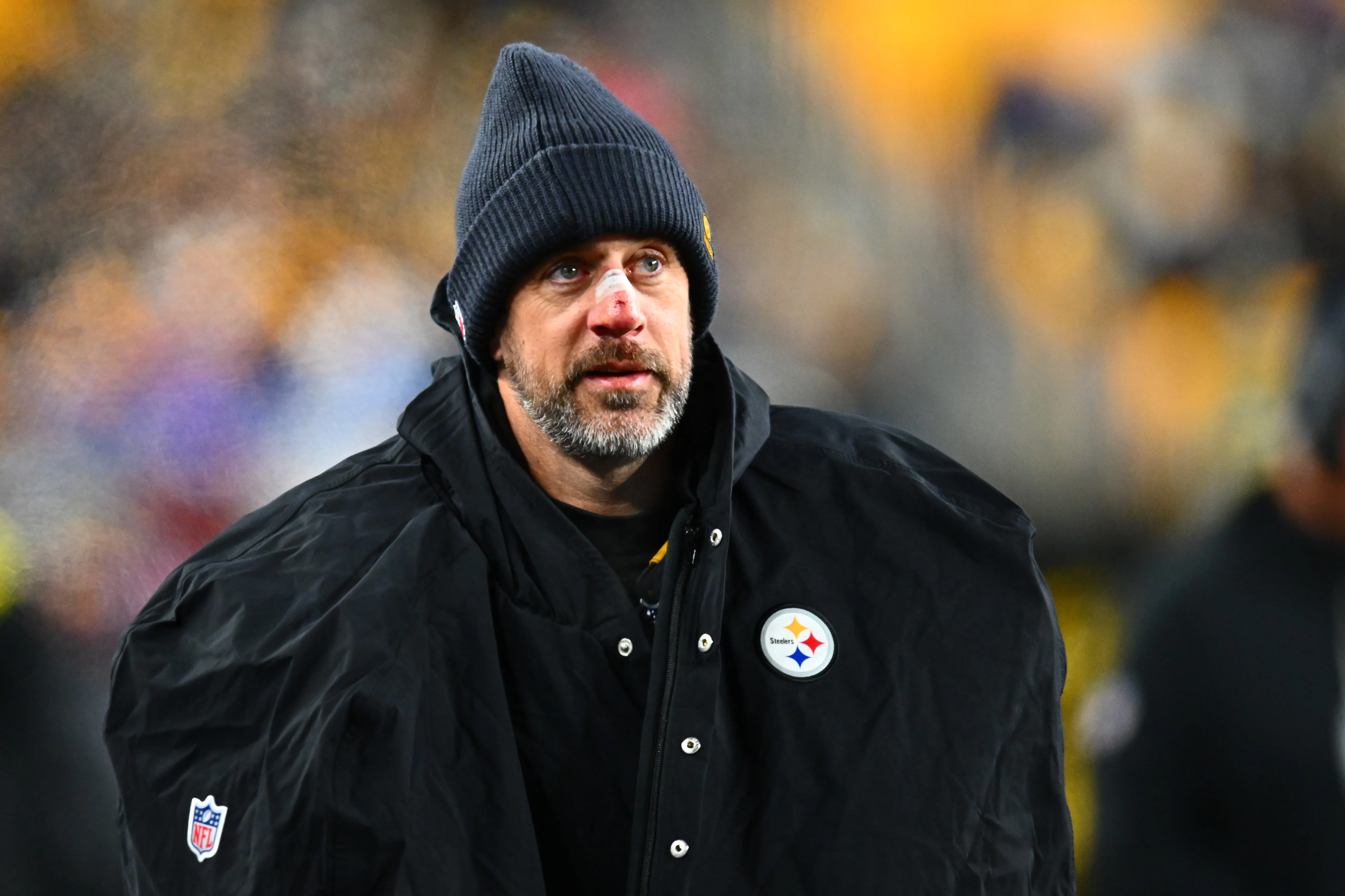 Aaron Rodgers wearing a Pittsburgh Steelers jacket and a beanie, with a bandage on his nose.
