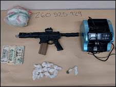 An assault rifle, cash, drugs, and a suspected explosive device on a table.