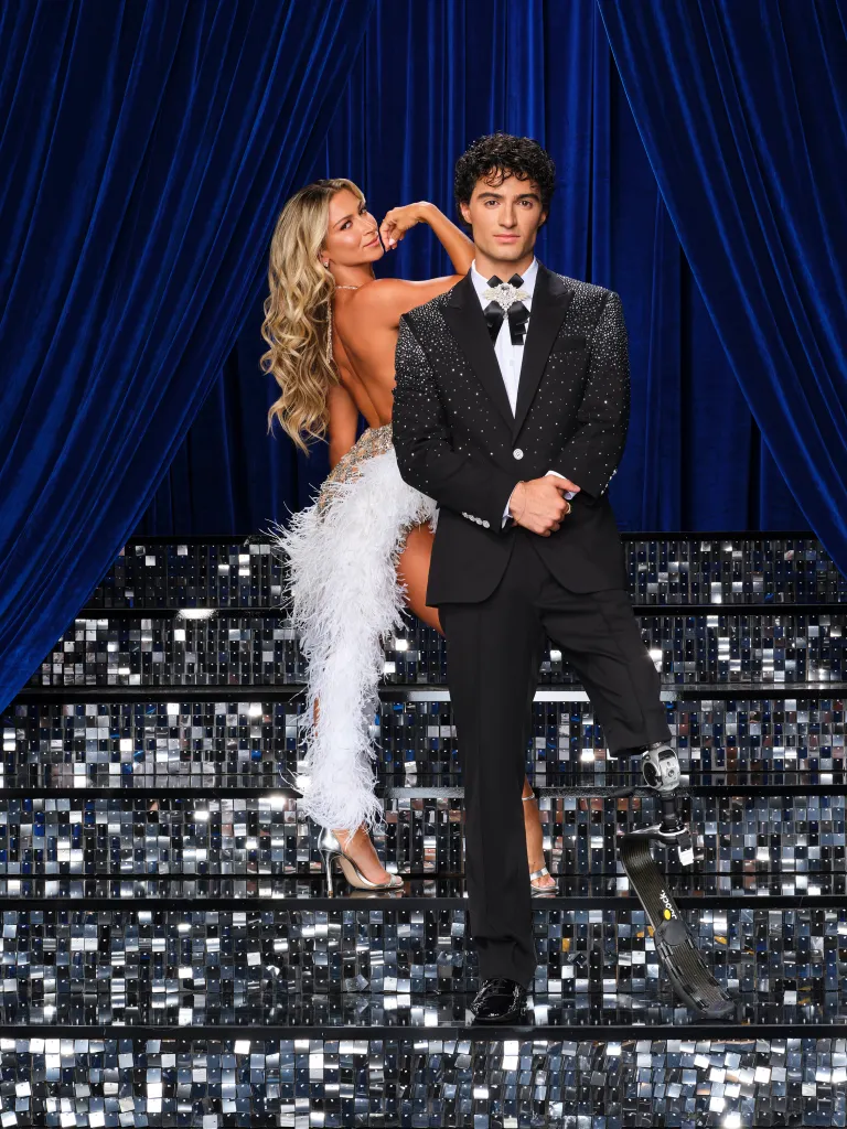 Daniella Karagach and Ezra Frech pose in formal wear on a set of sparkling stairs against a blue velvet curtain.