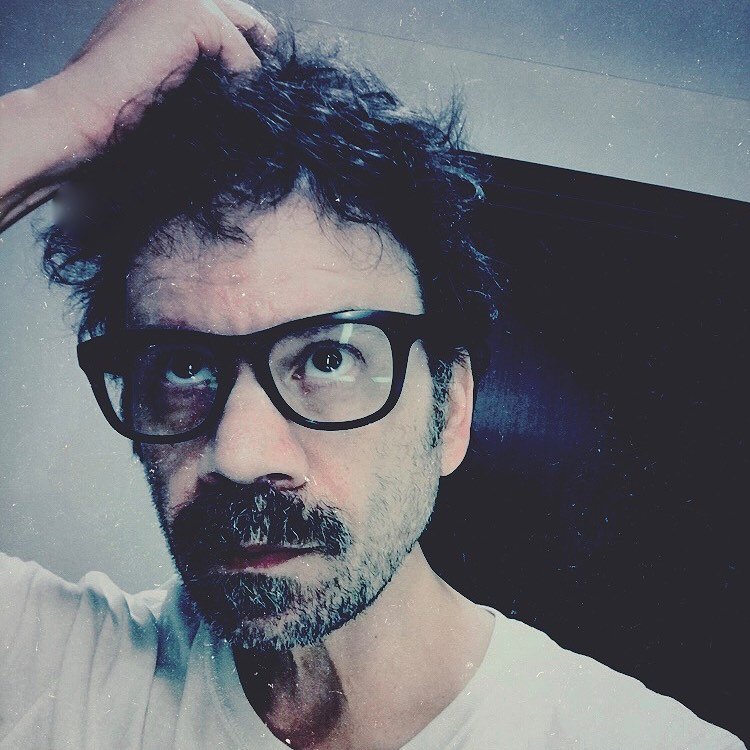 A man with dark, messy hair, glasses, and a mustache and beard, wearing a white t-shirt, looks up to the right while scratching his head.