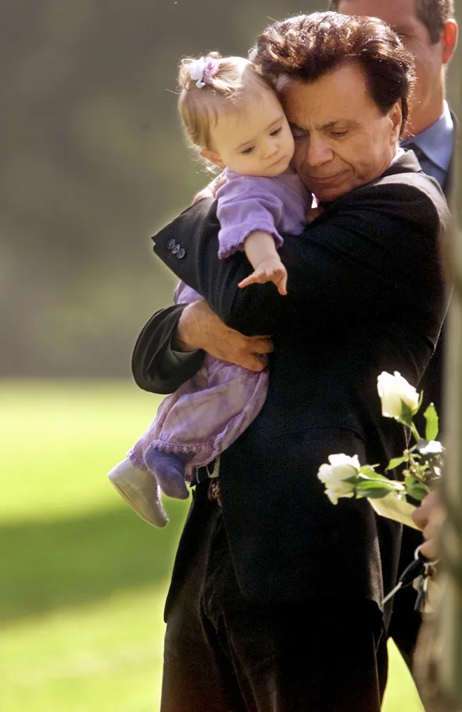 Robert Blake holds his 11-month-old daughter, Rose Lenore Sophie Blake, as she reaches for a white rose.