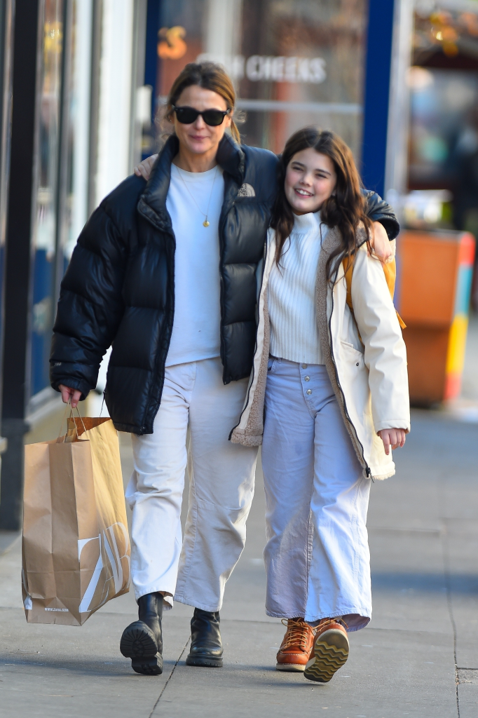 Keri Russell, in a black puffer jacket, white jeans and white top and black boots, and her daughter Willa Deary, in a cream jacket, cream top and white jeans, walk down a street, the actress with her arm around the child, both smiling.
