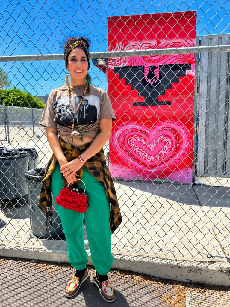 Adina Flores stands in front of a red mural featuring a black bird and a pink heart, behind a chain-link fence.