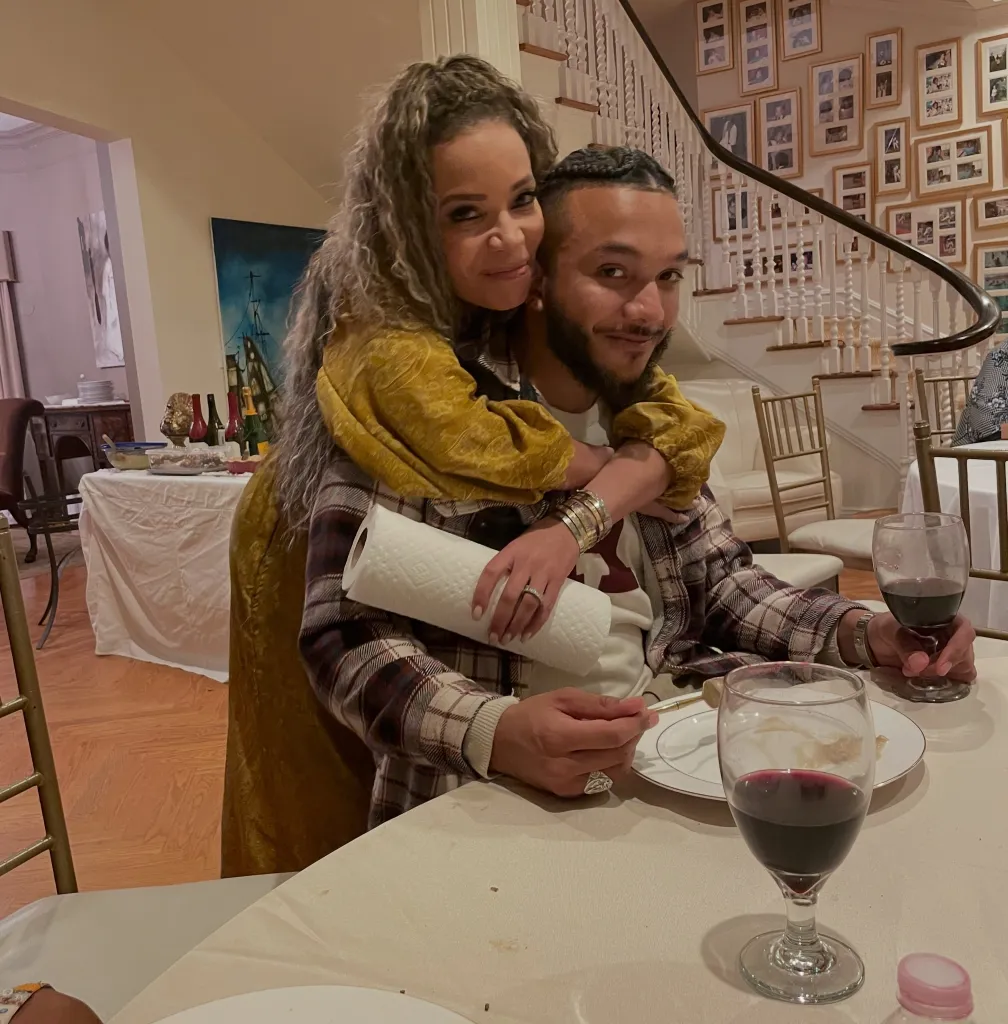 Sunny Hostin hugging her son Gabriel Hostin from behind at a table with plates and wine glasses.