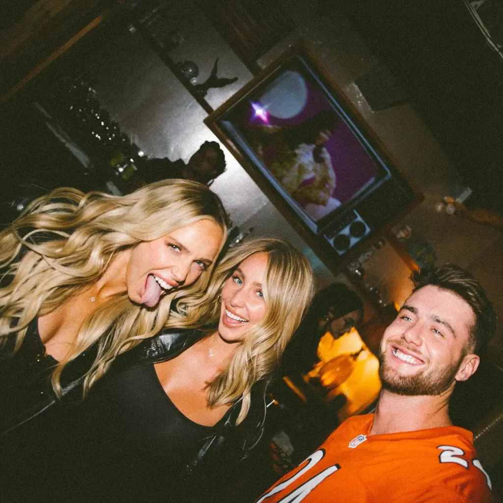 Alix Earle, Alex Cooper, and a man smiling together in a dimly lit setting.